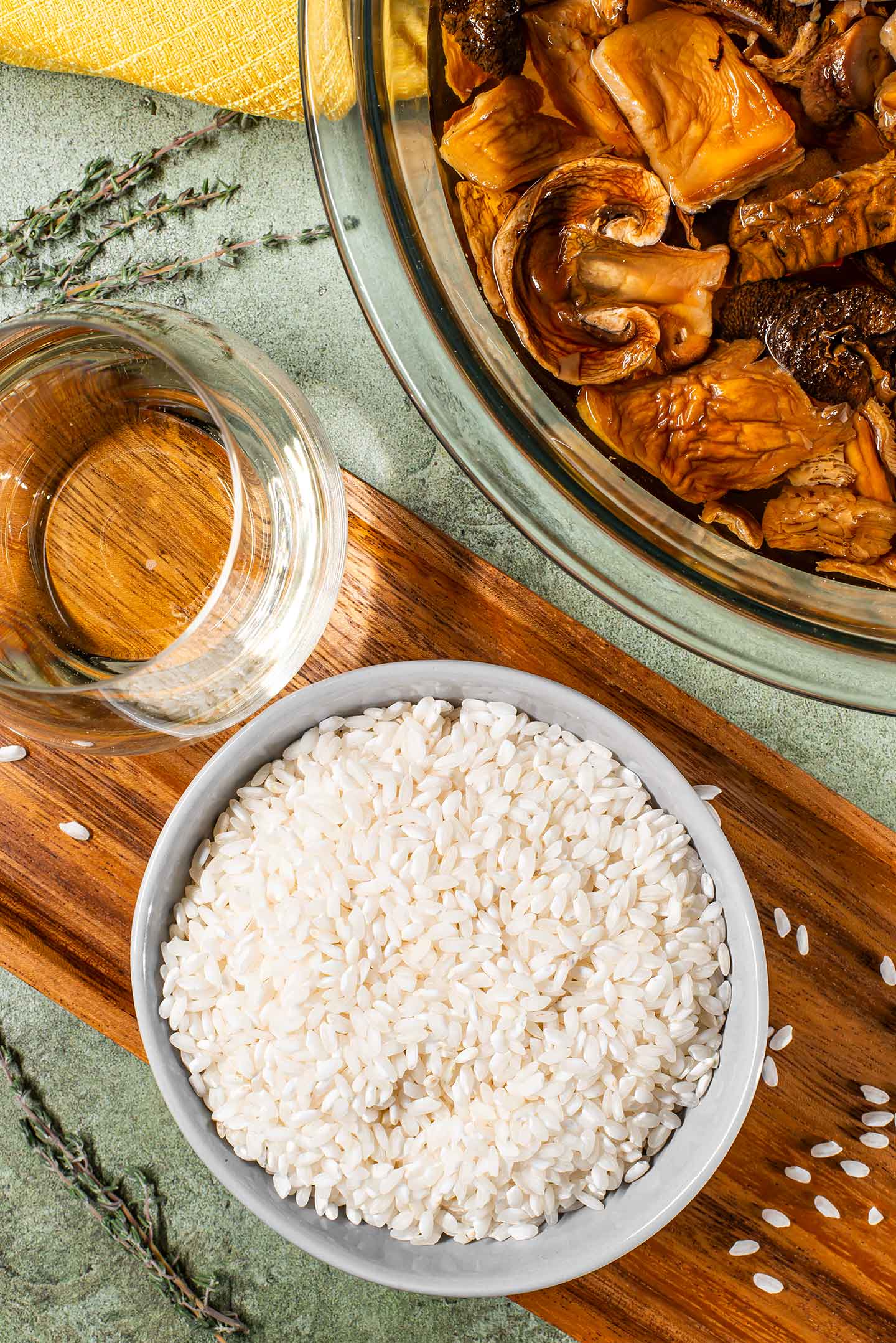 Top down view of arborio rice in a small dish with a glass of white wine nearby and a bowl of dried wild mushrooms rehydrating in water.