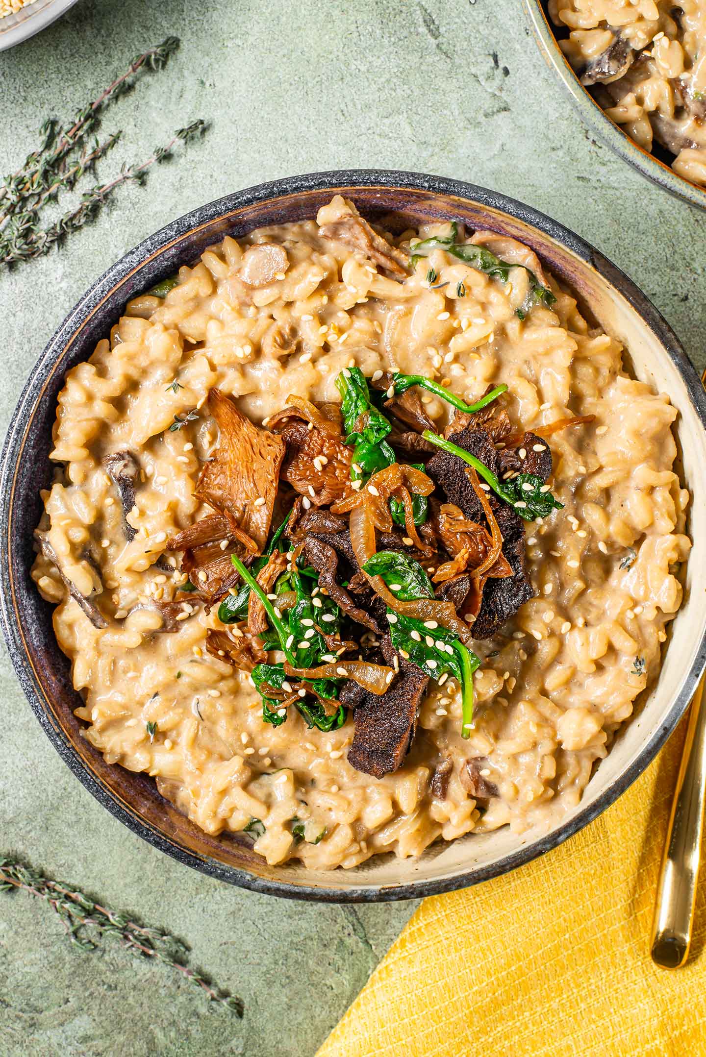 Top down view of creamy risotto in a dish topped with wild mushrooms, caramelized onion, and wilted spinach. Toasted sesame seeds and fresh thyme garnish.