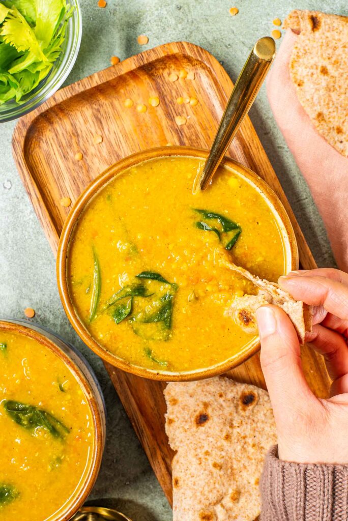 Top down view of a hand dipping roti into a golden red lentil curried vegetable soup.