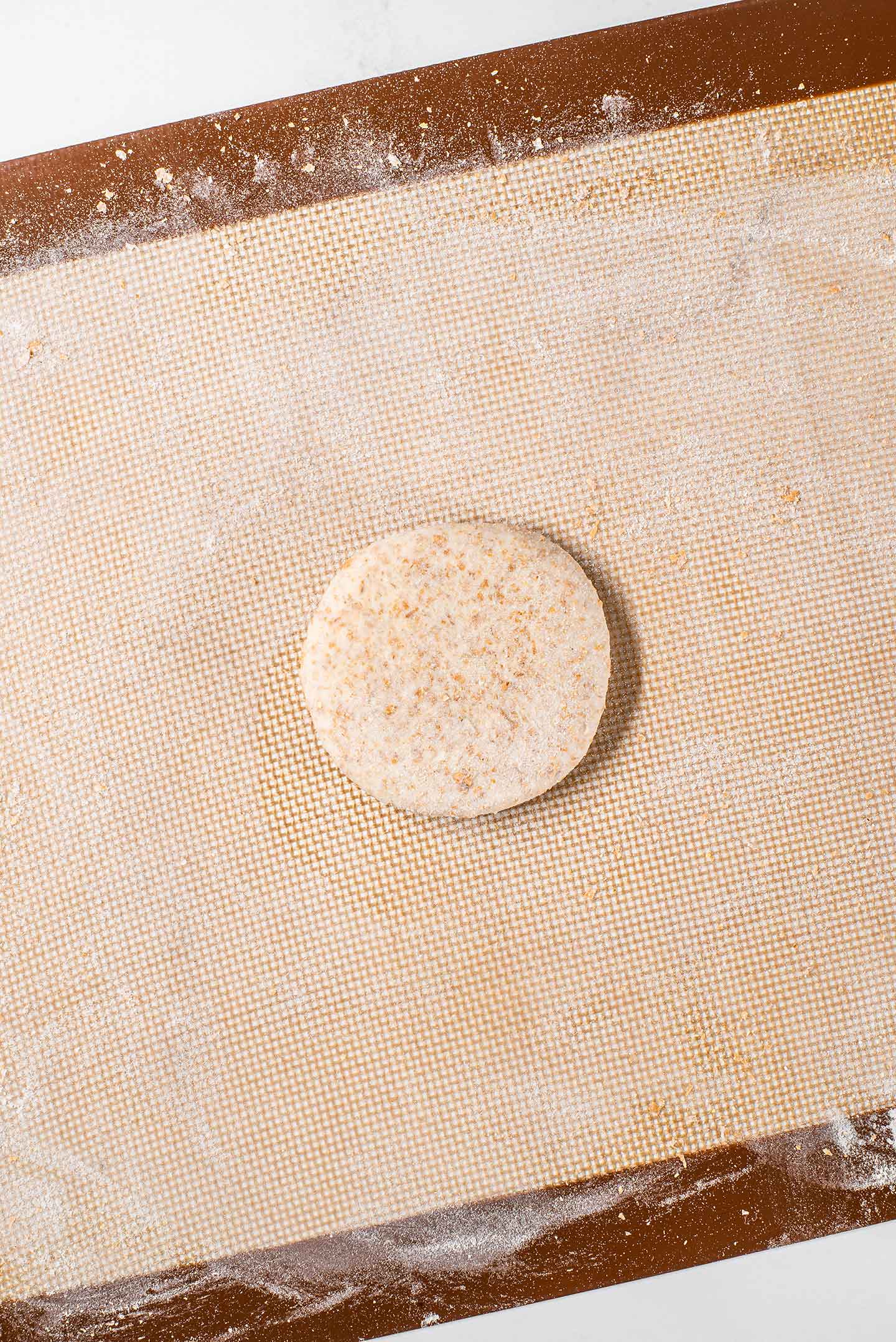 Top down view of a single flattened ball of roti dough on a silicone mat before it is rolled into a larger flat roti.
