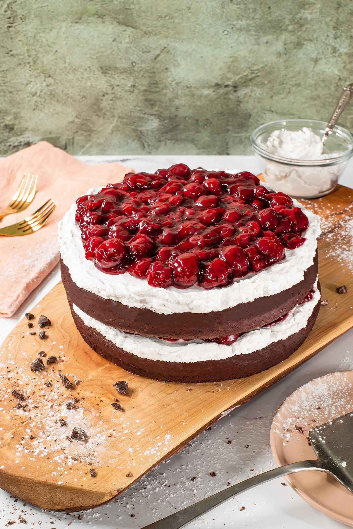 Side view of a vegan black forest cake topped with coconut whipped cream and a thick cherry sauce. The sides of the cake are bare, showing off the two layers of chocolate cake with more whip and cherries in the centre.