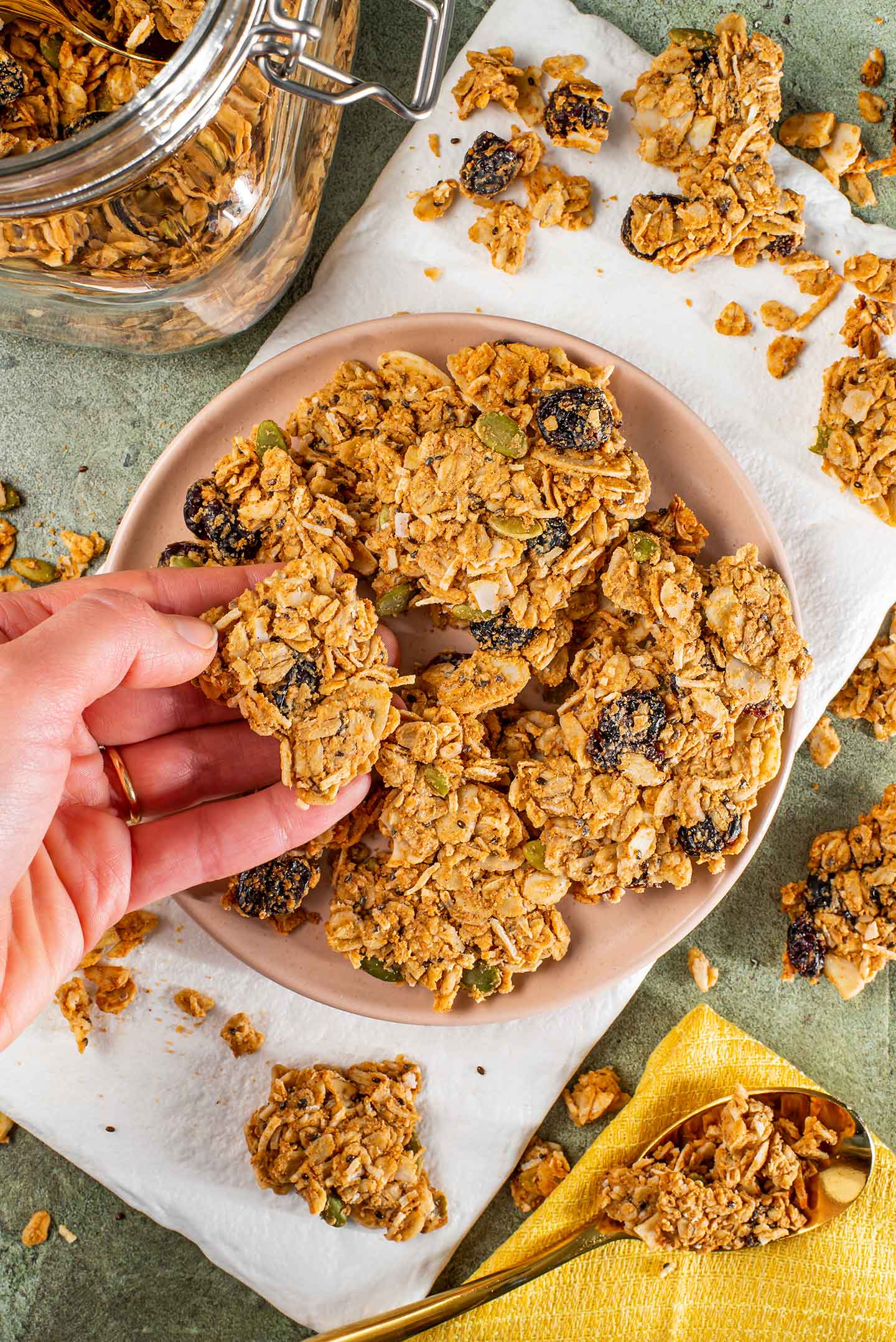 Top down view of a hand holding a piece of chunky peanut butter granola to show the size of the granola clusters displayed on a plate below. 