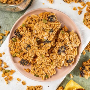 Top down view of crunchy peanut butter granola clusters on a plate. The granola is speckled with pumpkin seeds, sliced almonds, and dried cranberries.