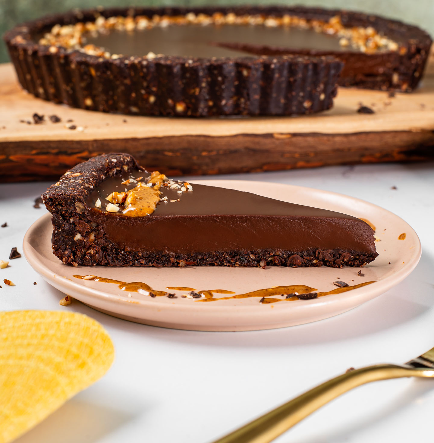 A slice of vegan chocolate almond tart sits on a plate. The chocolate ganache filling is soft but fully set. The crust holds firm and the slice is decorated with an almond butter drizzle.