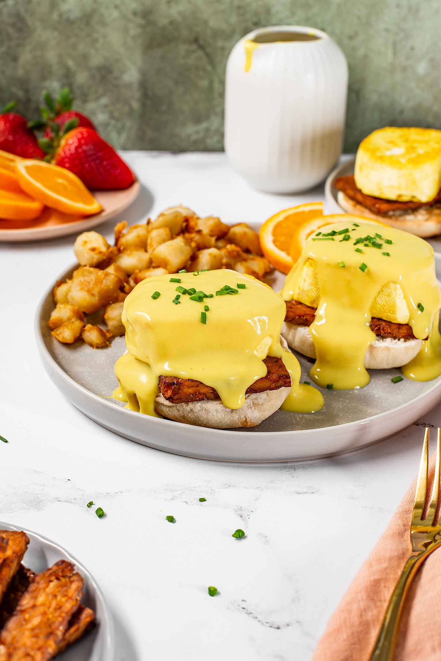 Side view of vegan eggs benedict on a plate with vegan home fries and orange slices. Two tofu eggs covered in creamy vegan hollandaise sauce sit atop tempeh bacon on english muffins.