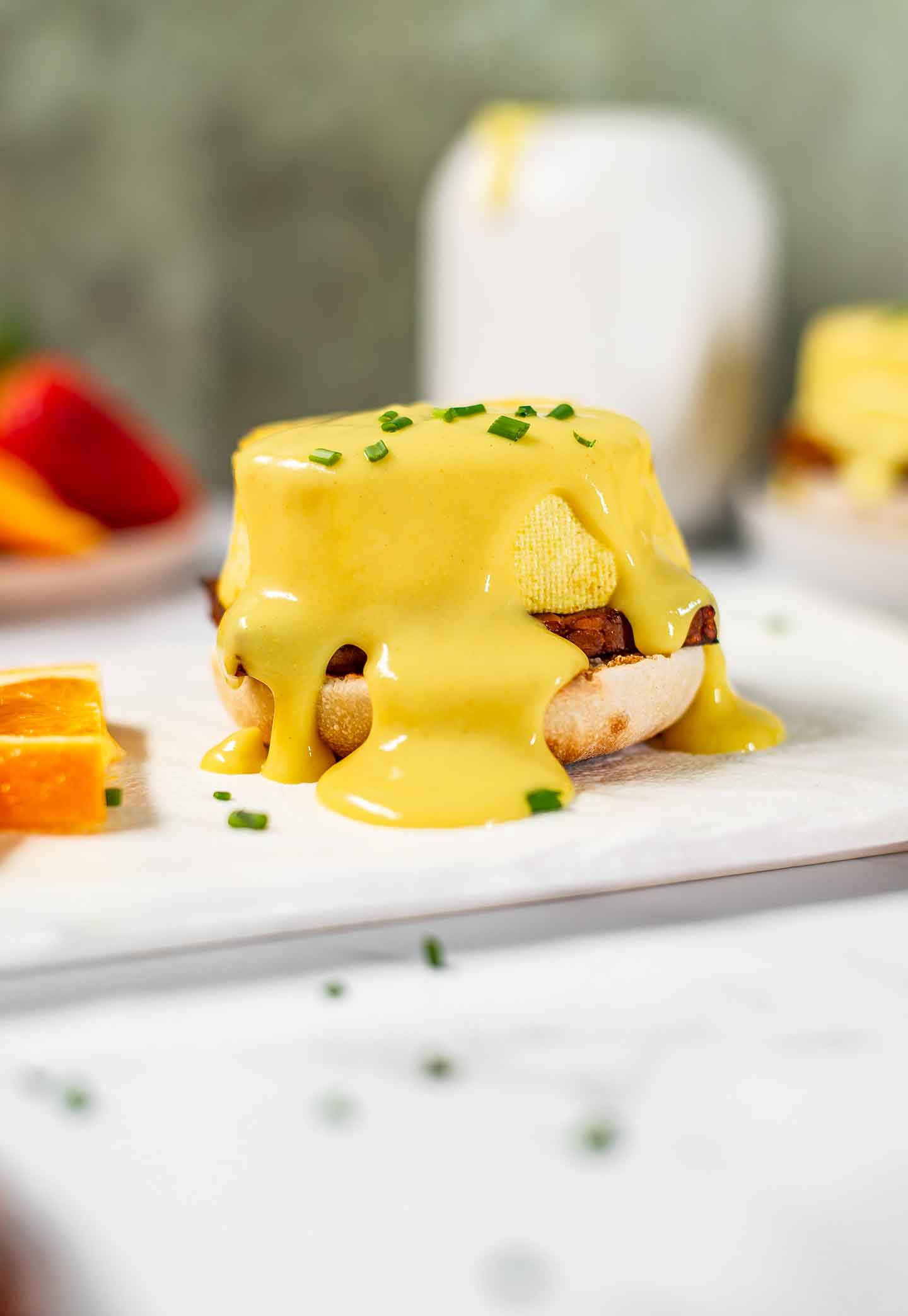Thick vegan hollandaise sauce, tinged yellow, runs over top and down the sides of a vegan eggs benedict.