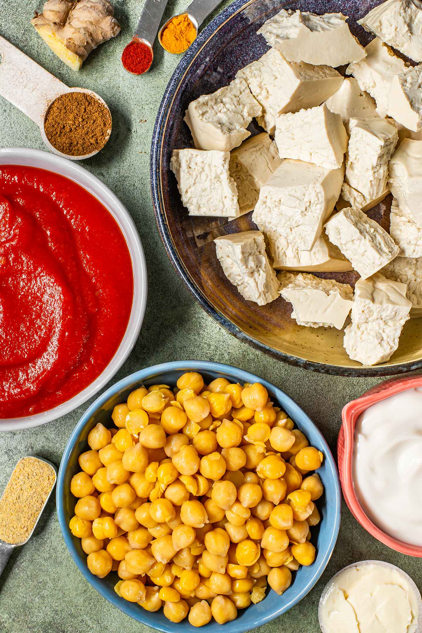 Top down view of ingredients. Hand pulled chunks of firm tofu, chickpeas, crushed tomatoes, vegan yogurt, and spices.