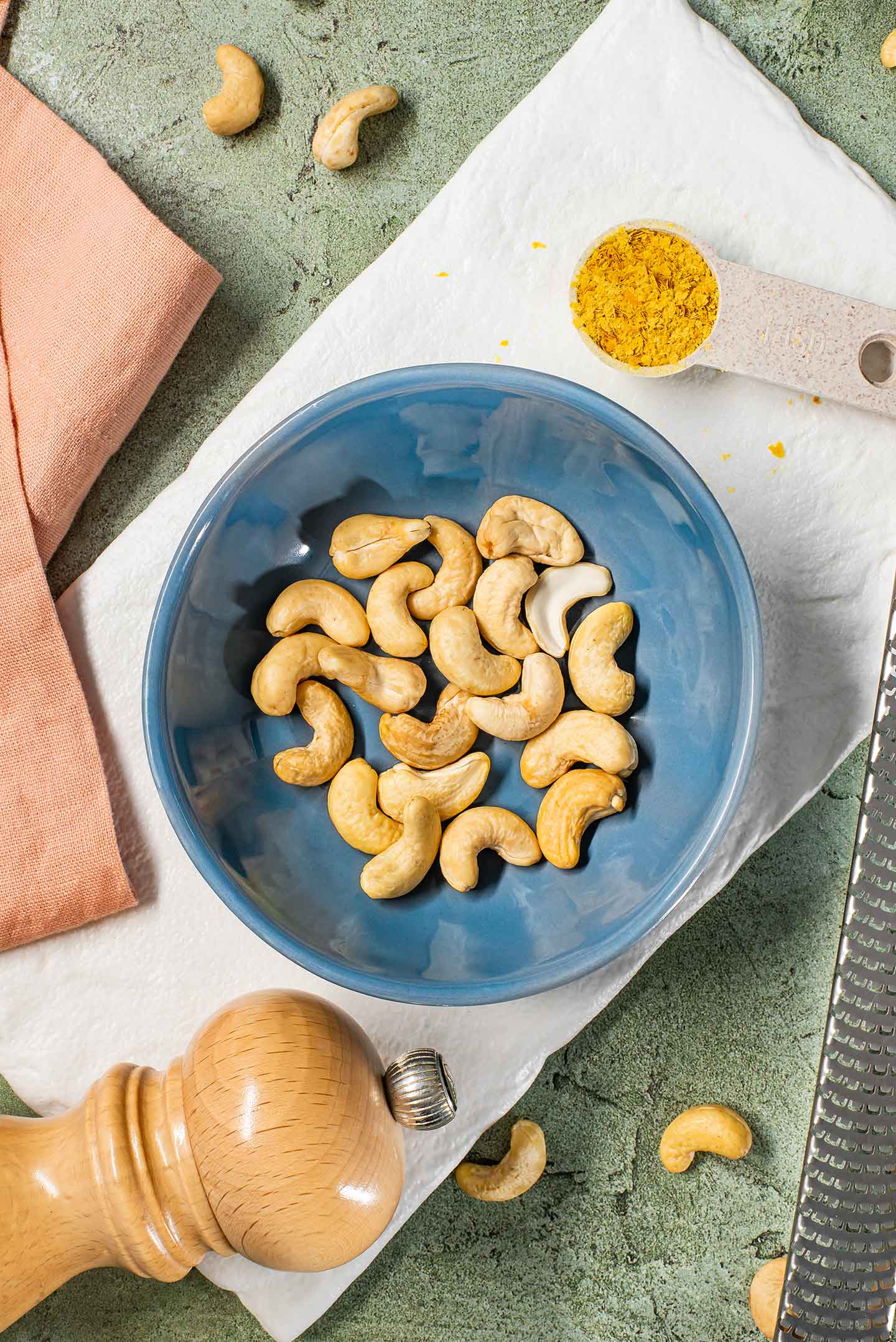 Top down view of cashews in a small bowl with salt and nutritional yeast resting on a white tray.