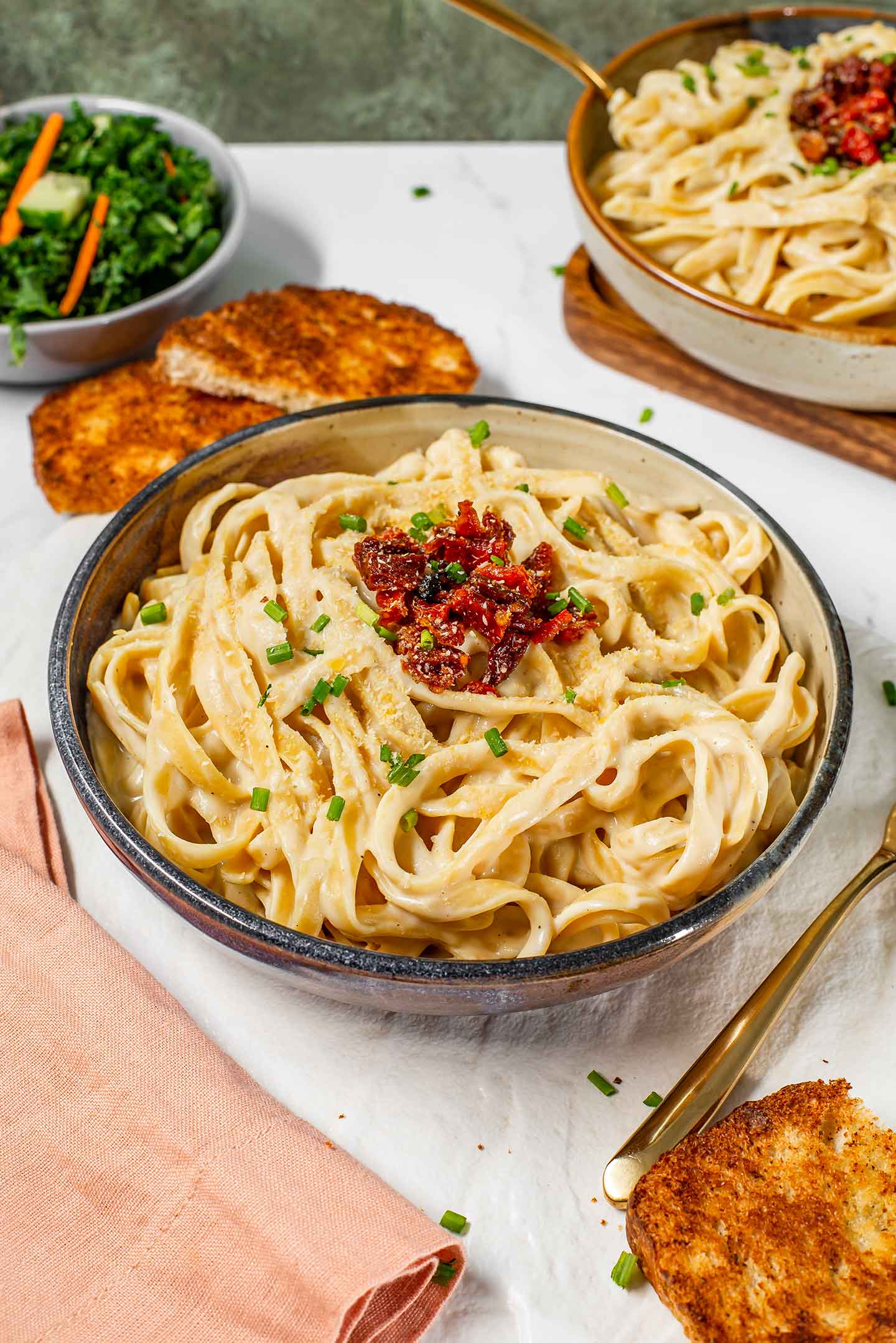 Side view of creamy dairy-free fettuccine alfredo in a pasta bowl. The sauce is smooth and the pasta is topped with sun-dried tomatoes, chives, and vegan parmesan.