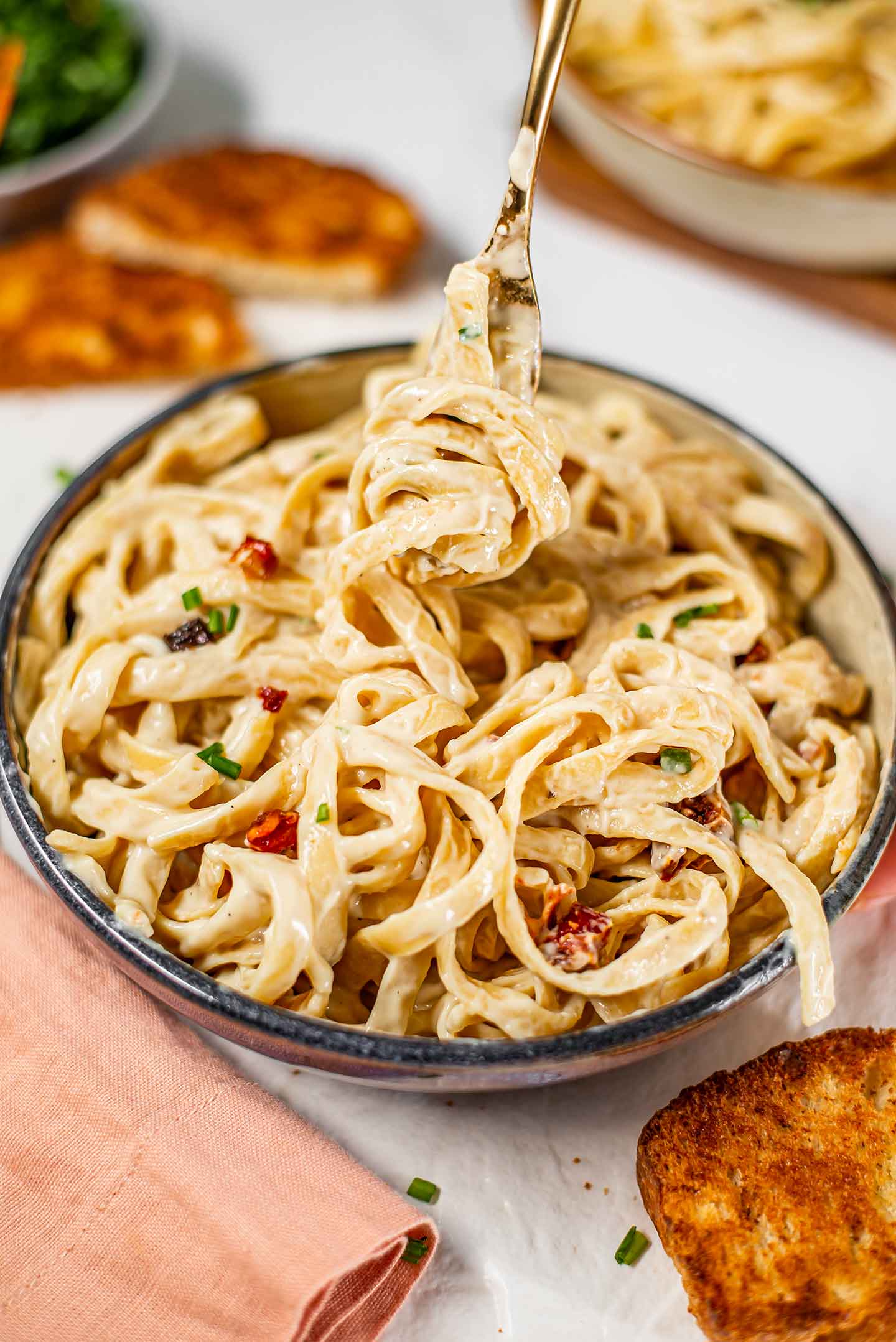 Fettuccine noodles coated in a creamy vegan alfredo sauce wrap around a fork lifting up from a bowl of pasta.