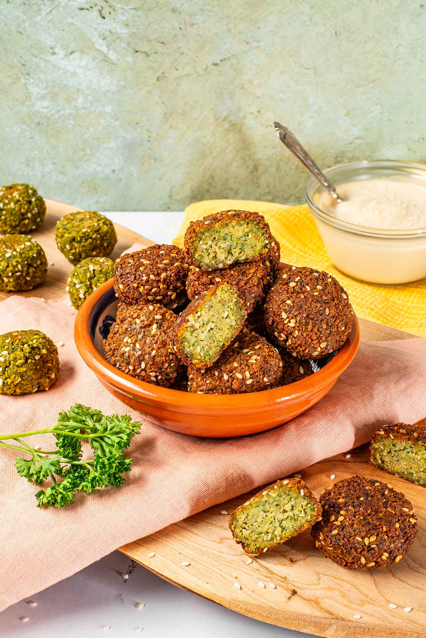 Side view of crispy falafel piled in a small dish. The outside of the falafel is dark brown from frying but the inside is green and fluffy. Some baked falafel are green on the outside an spread on a tray in the background.