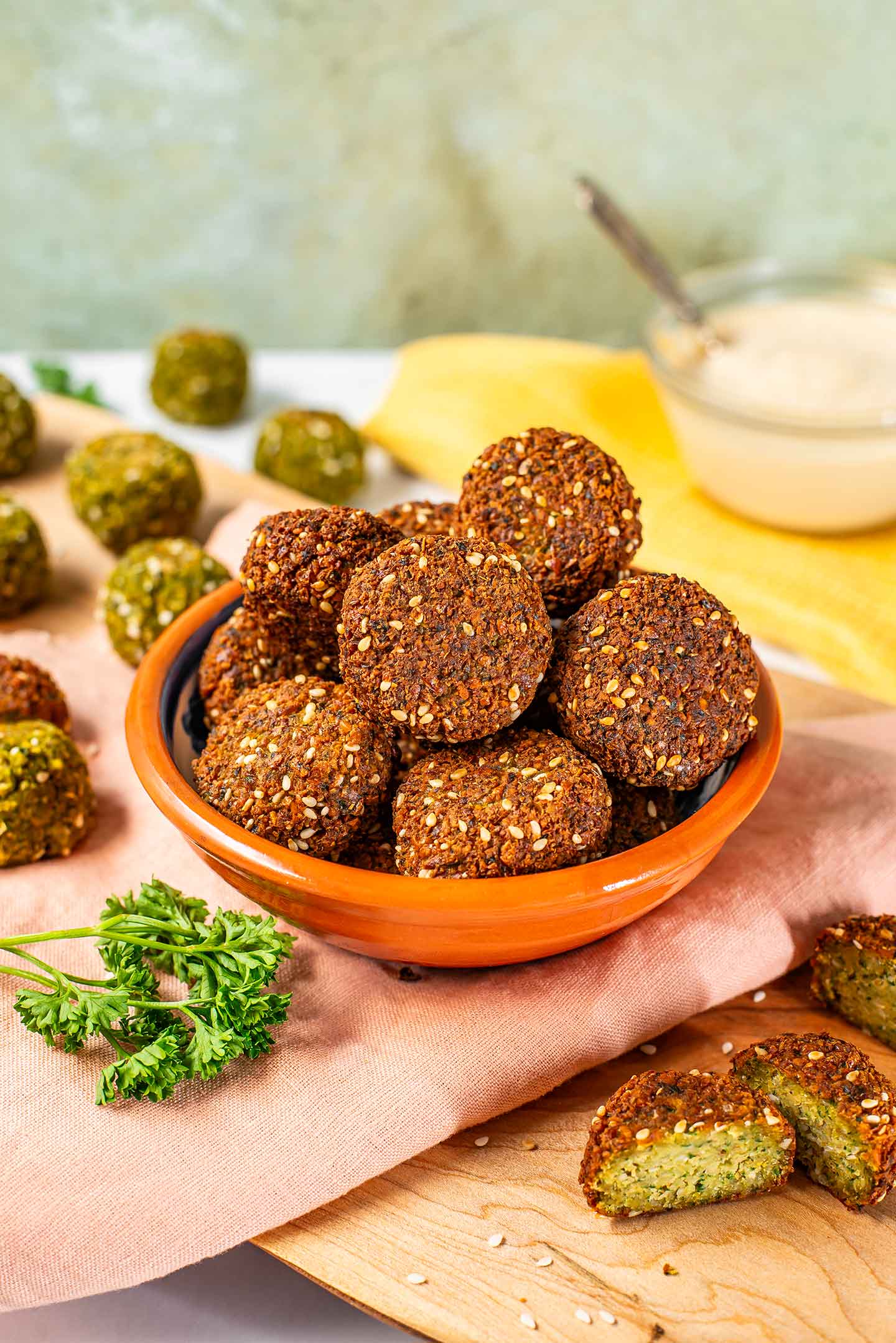 Side view of perfectly crispy falafel in a small dish. A tahini dressing, baked falafel, and fresh sprigs of parsley surround the bowl.