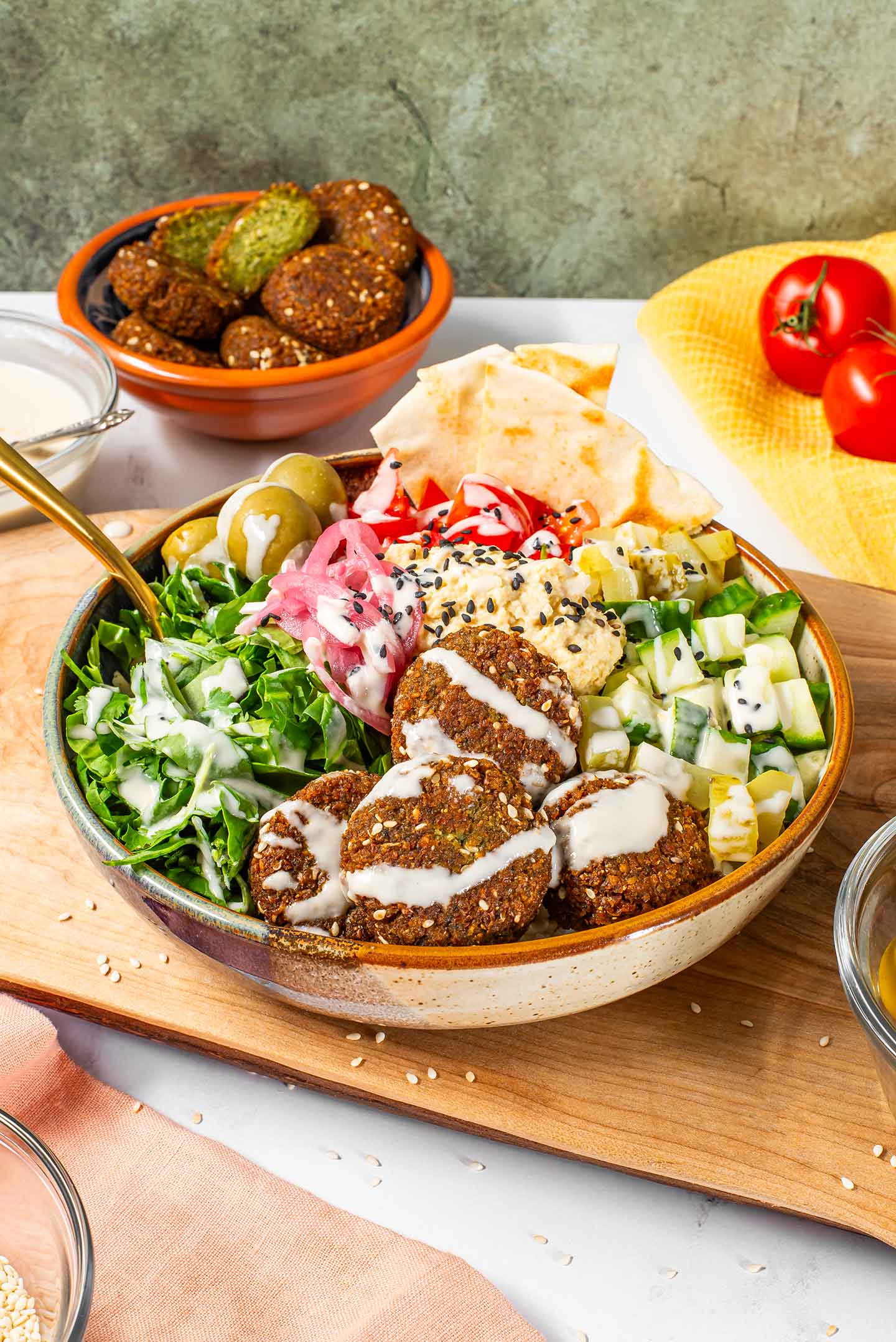 A colourful falafel bowl consisting of crispy fried falafel, cucumber, tomato, greens, olives, pickled red onion and drizzled with a creamy tahini dressing. Toasted pita rests in the bowl and extra falafel are in the background.
