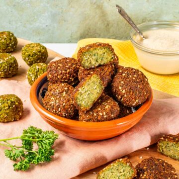 Side view of crispy falafel piled in a small dish. The outside of the falafel is dark brown from frying but the inside is green and fluffy. Some baked falafel are in the background.
