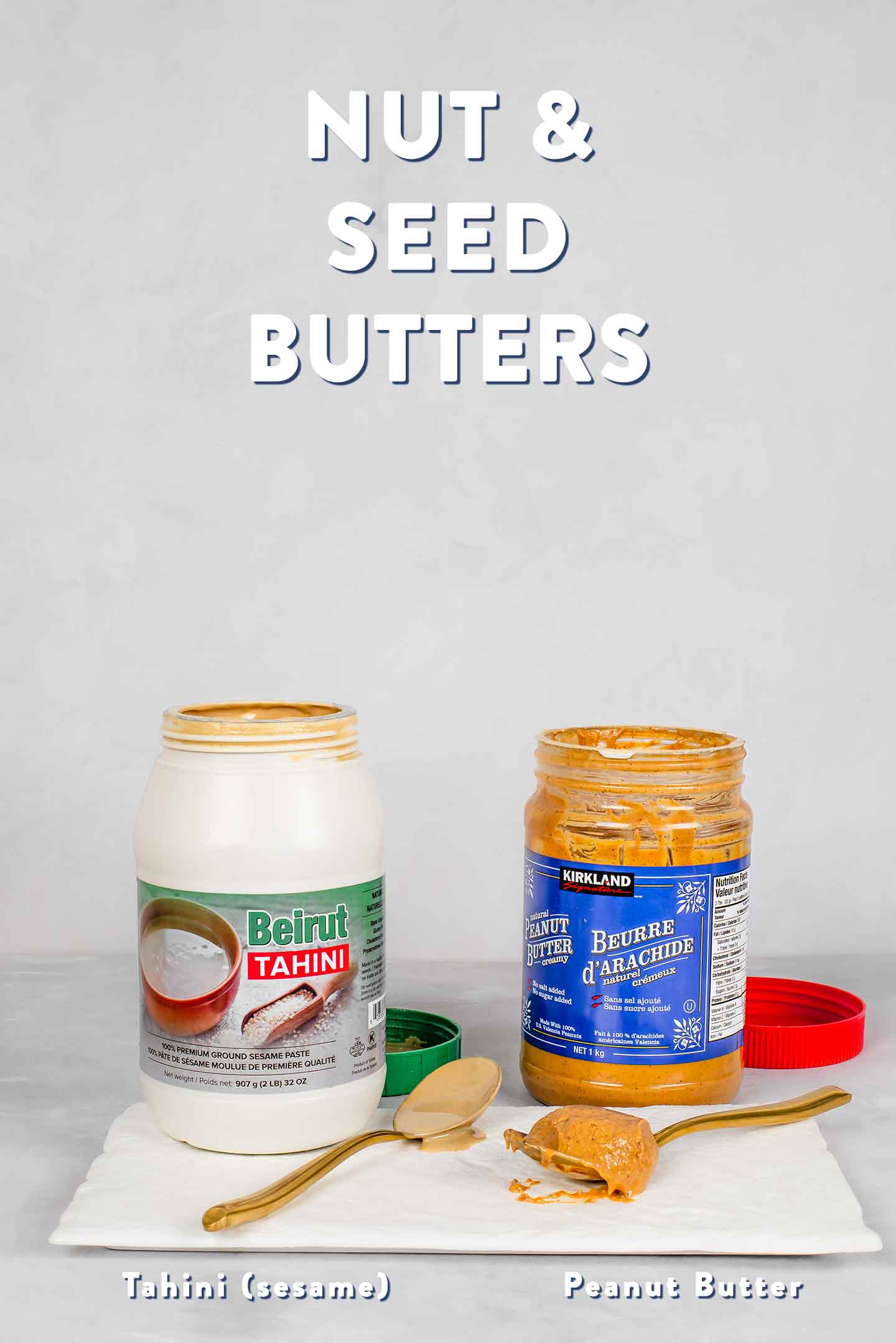 A jar of tahini and a jar of natural peanut butter sit with their lids off. Spoons with the two butters lay on a tray in front of the jars.