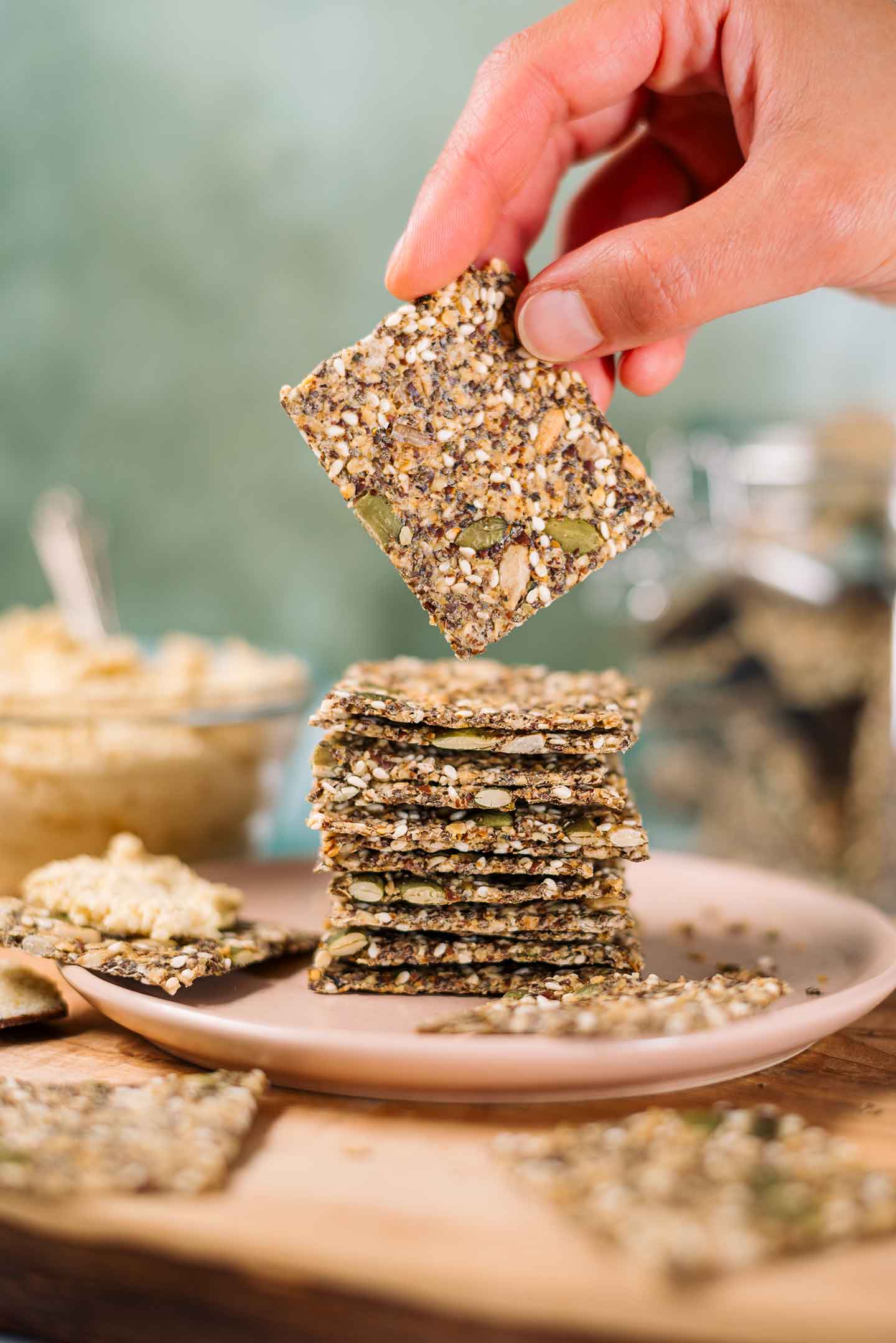 A hand holds a gluten-free seed cracker up from a stack of thin and crispy crackers made entirely from a combination of seeds. A jar of crackers and a bowl of hummus are in the background.