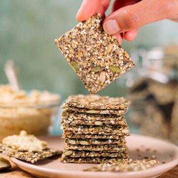 A hand holds a gluten-free seed cracker up from a stack of thin and crispy crackers made entirely from a combination of seeds.