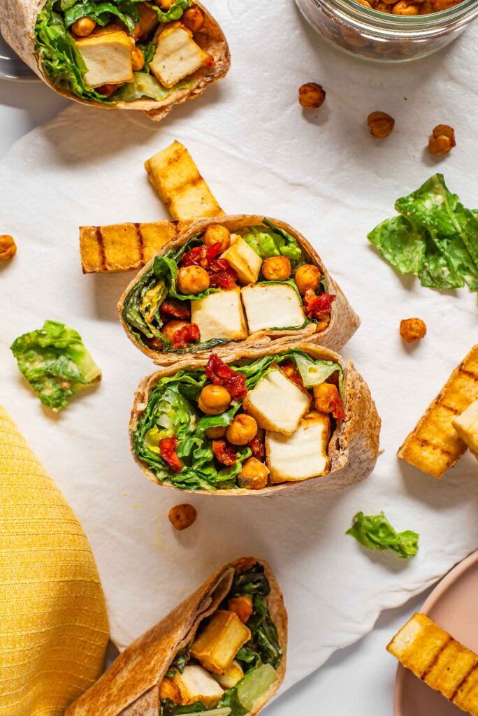 Top down view of two vegan caesar wraps cut in half to show the strips of grilled tofu, romaine lettuce with vegan caesar dressing, roasted chickpeas, and sun-dried tomato "bacon bits".