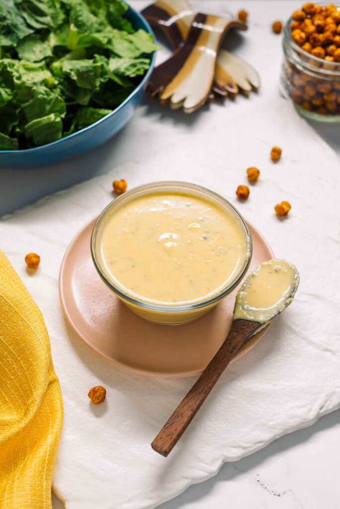 Side view of a creamy dairy-free caesar dressing in a small bowl. A spoon covered with dressing rests beside the bowl. A bowl of romaine lettuce fills the background.