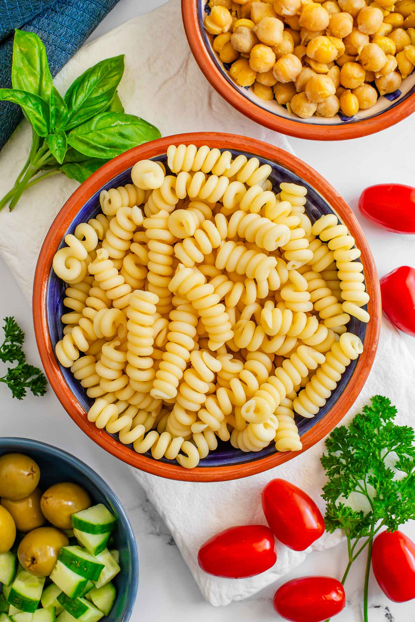Top down view of ingredients for pasta salad in a jar. Fusilli pasta, olives, grape tomatoes, chopped cucumber and fresh herbs rest on a tray.