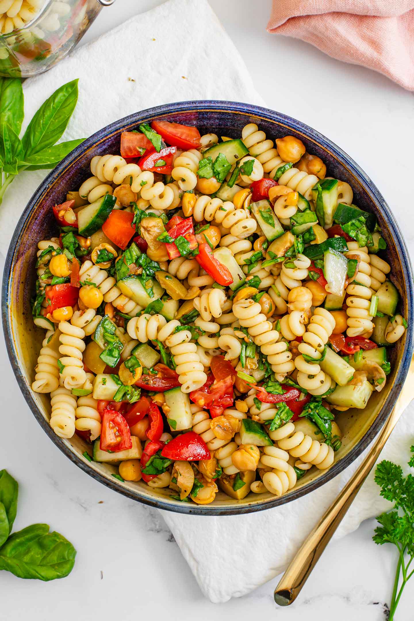 Top down view of a vegan pasta salad in a bowl. Chopped tomatoes, cucumbers, olives, spinach, and herbs are mixed with fusilli noodles and chickpeas.