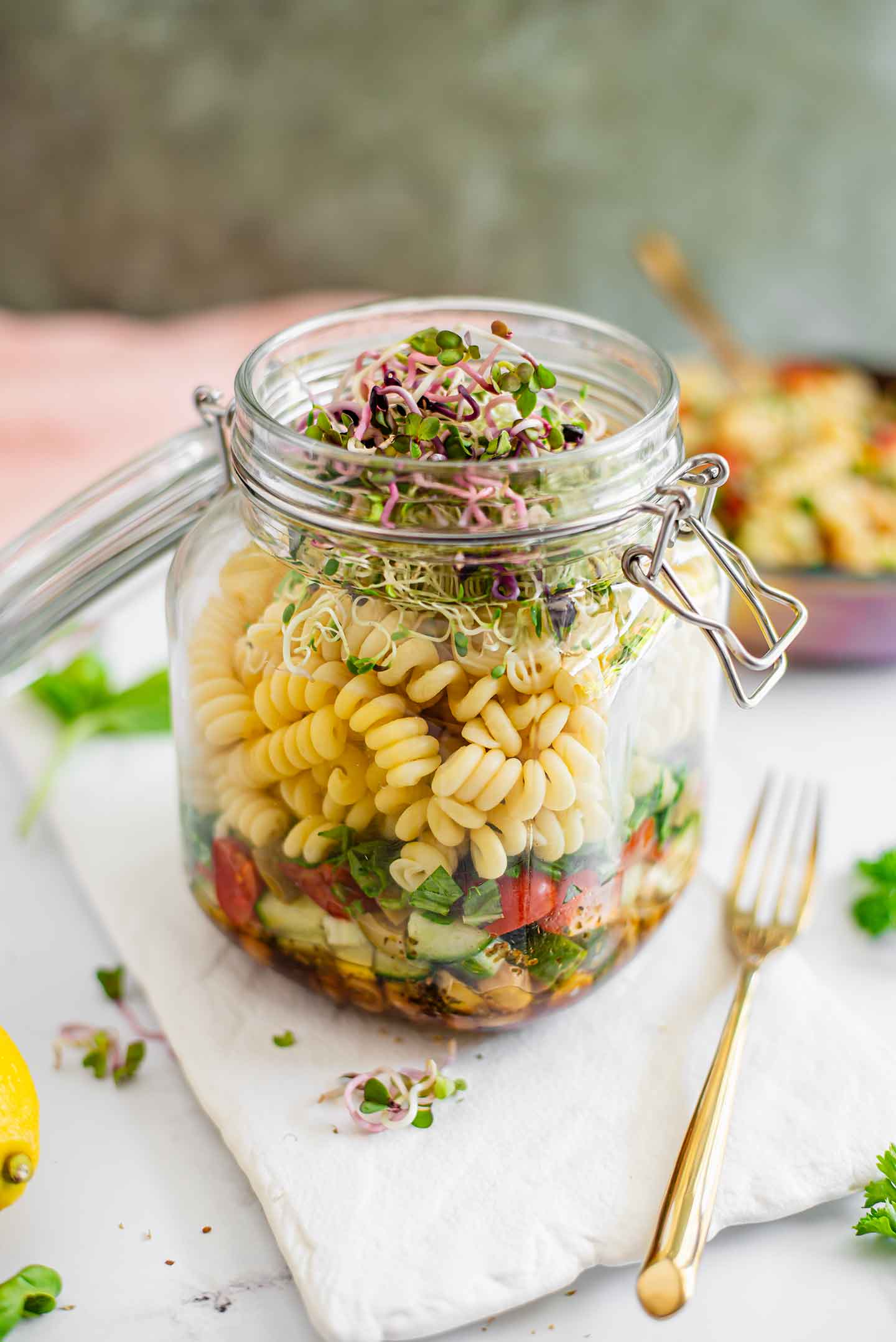 Side view of a layered pasta salad prepared in a glass jar. Dressing, vegetables, and chickpeas are on the bottom while fusilli pasta is layered on top. Colourful radish sprouts garnish the top.