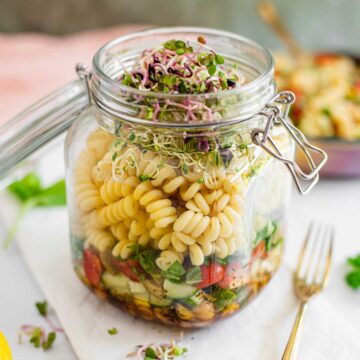Side view of a layered pasta salad prepared in a glass jar. Dressing, vegetables, and chickpeas are on the bottom while fusilli pasta is layered on top.