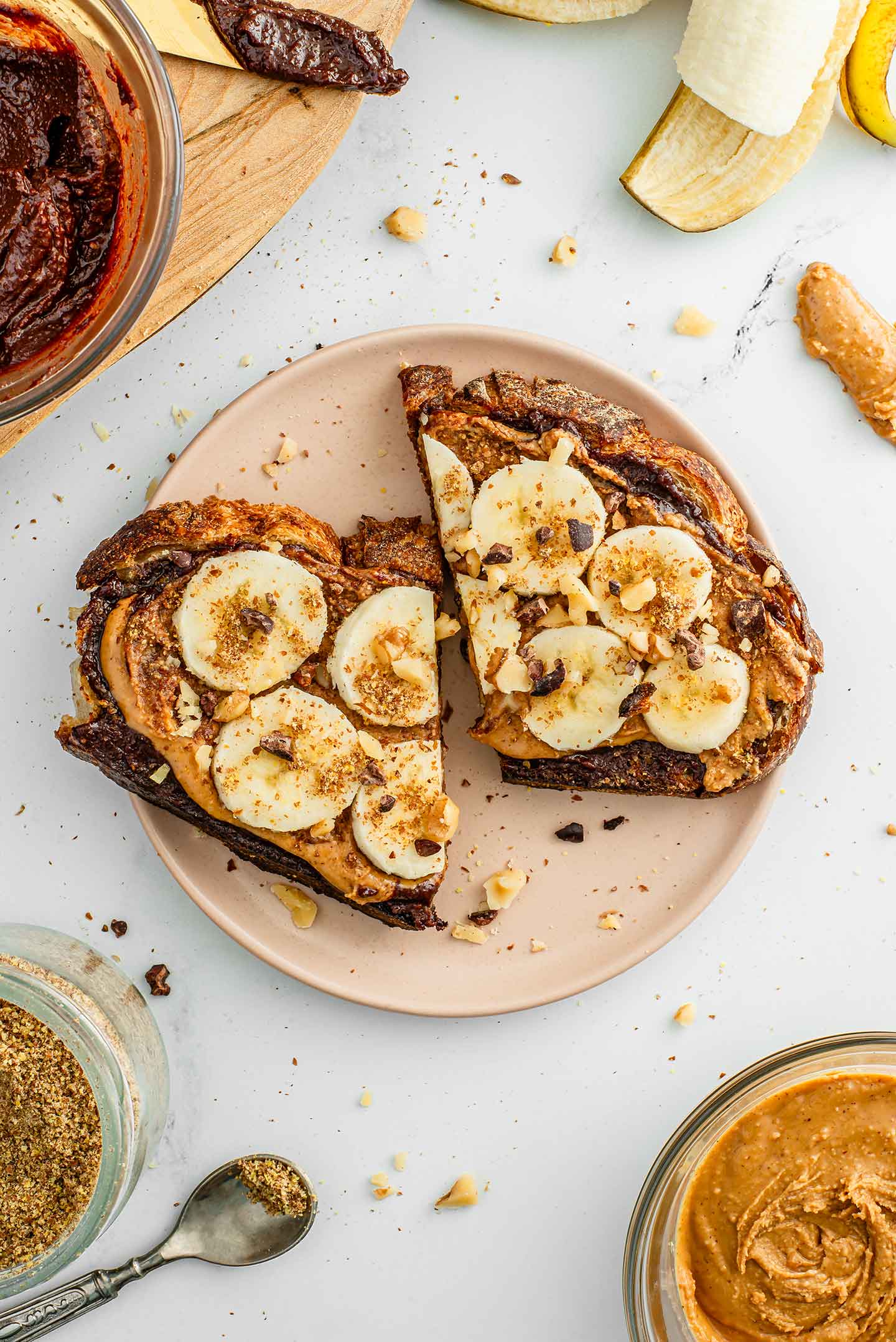 Top down view of an open-faced vegan Nutella sandwich on a plate. It is topped with peanut butter, sliced banana, ground flax seed, cacao nibs, and toasted chopped walnuts. Extra ingredients surround the plate.