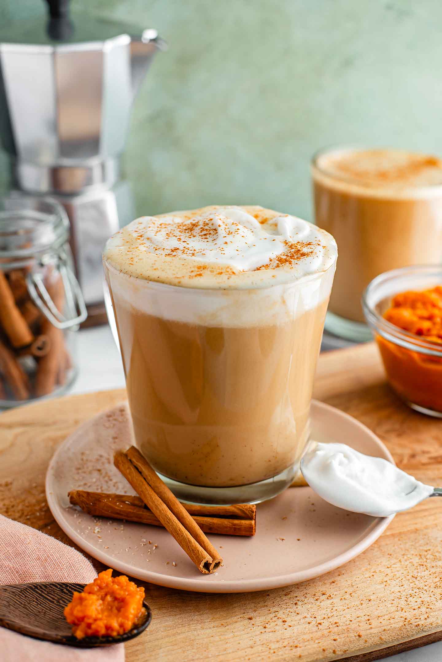 Side view of a warm vegan pumpkin spice latte in a glass mug. Whipped cream garnishes the top and a second latte with no whip is in the background.