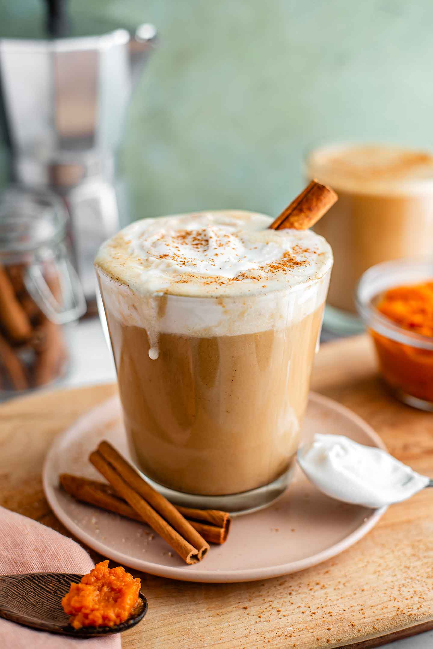 Dairy-free pumpkin spice latte topped with coconut whipped cream fills a glass mug. The whip is dusted with cinnamon and dribbles slightly down the side of the mug. A cinnamon stick rests in the festive latte.