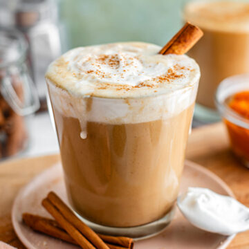 Dairy-free pumpkin spice latte topped with coconut whipped cream fills a glass mug. The whip is dusted with cinnamon and dribbles slightly down the side of the mug.
