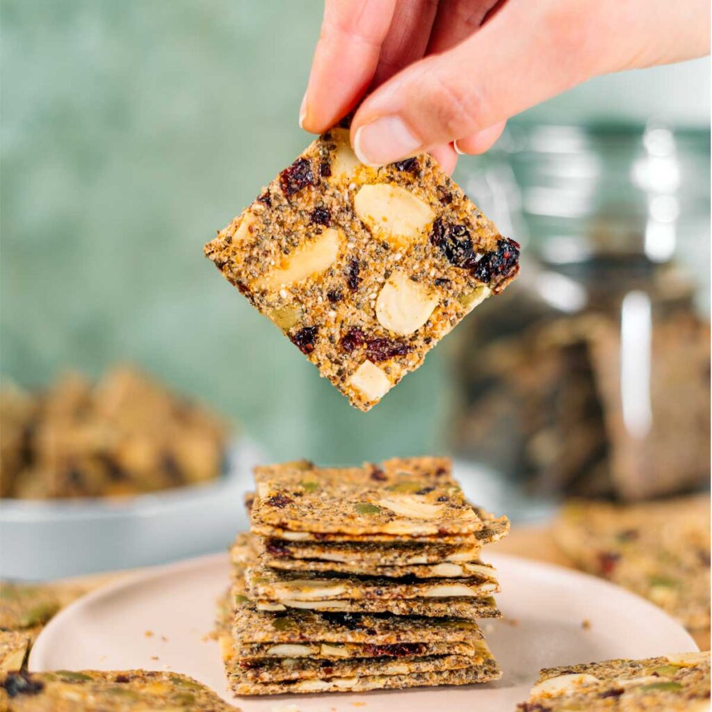 Side view of a hand holding one seed cracker above a stack of thin crackers. The crackers are golden and speckled with sliced almonds, pumpkin seeds, and dried cranberries.