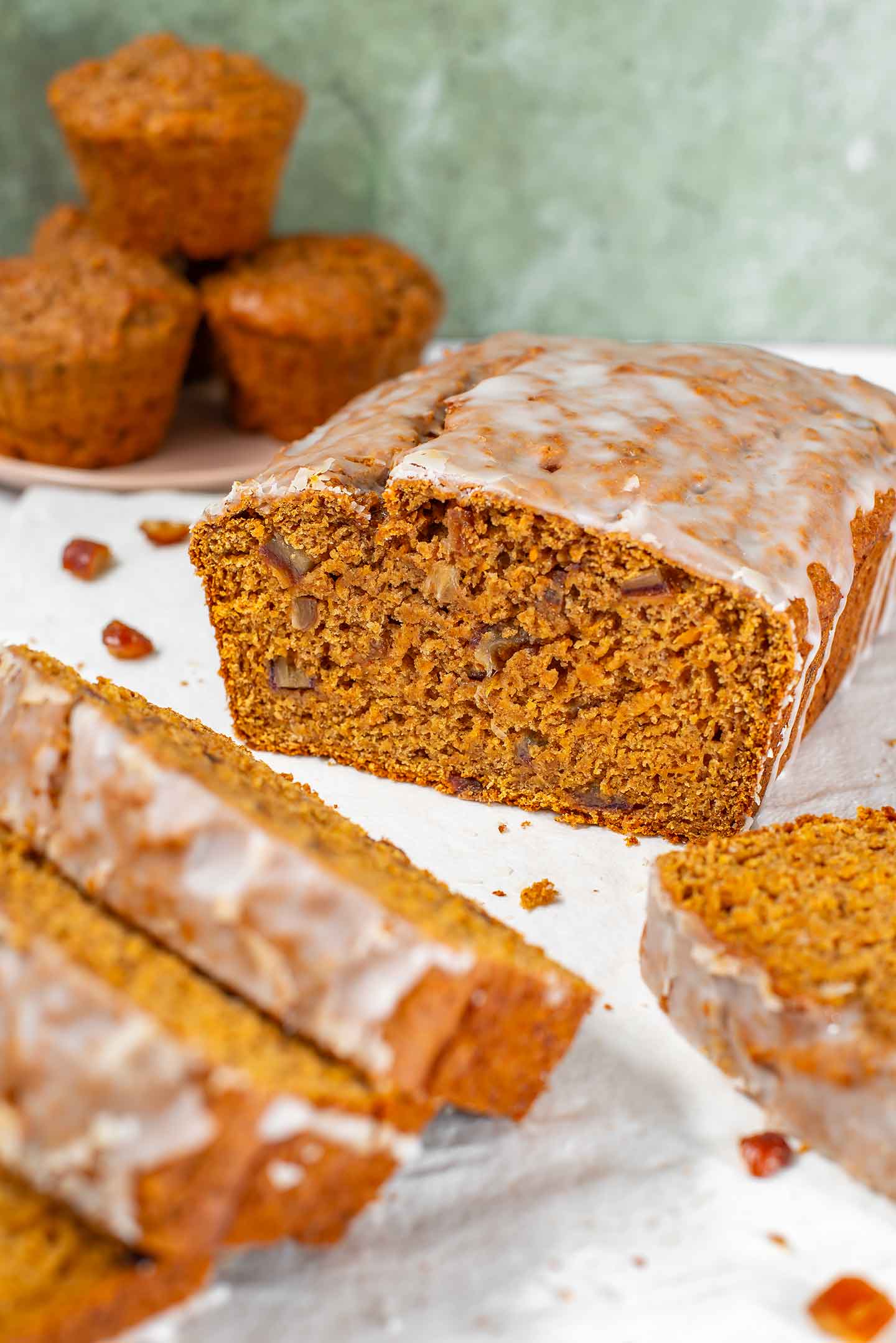 Side view of a glazed loaf of vegan sweet potato bread. The loaf has been cut exposing the tender crumb speckled with chopped dates. Muffins are stacked on a plate in the background.