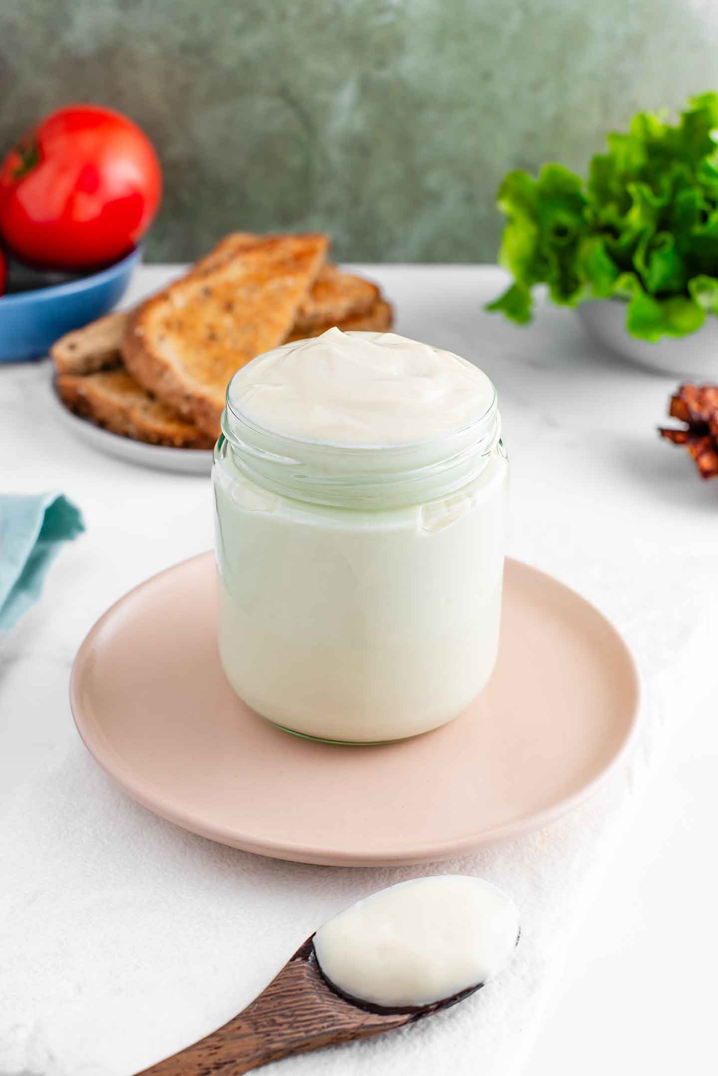 Side view of oil-free mayonnaise filling a jar. The mayo is thick and creamy white in colour. A spoonful of thick mayo rests in front of the jar and sandwich ingredients fill the background.