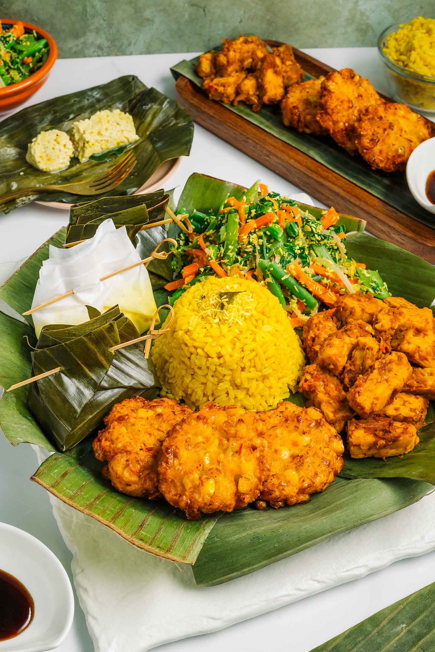A tasting platter of Balinese food presented on banana leaves. A mound of turmeric rice sits in the centre with tofu pepes, urab coconut and vegetable salad, tempeh, and corn fritters surrounding.