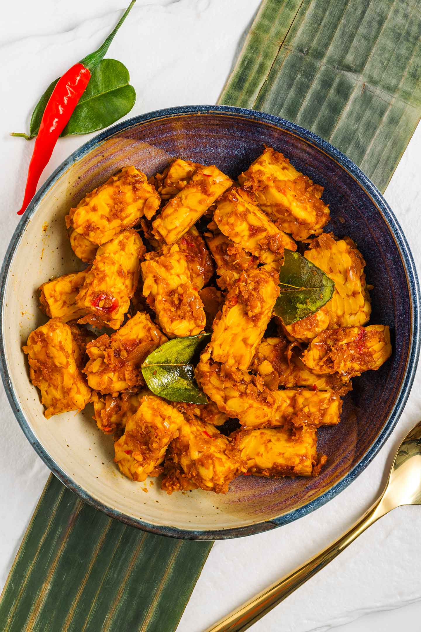 Vegan Balinese tempeh fills a shallow bowl. The tempeh is golden and spiced. Kaffir lime leaves garnish. A red chilli lays beside the bowl and a banana leaf lays underneath.