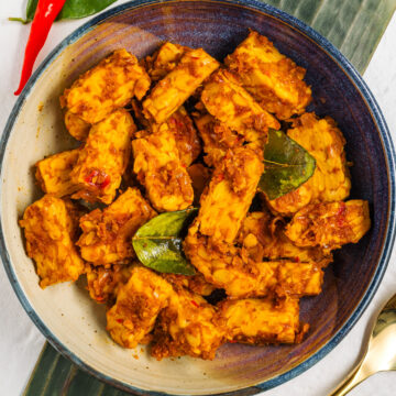 Vegan Balinese tempeh fills a shallow bowl. The tempeh is golden and spiced. Kaffir lime leaves garnish.