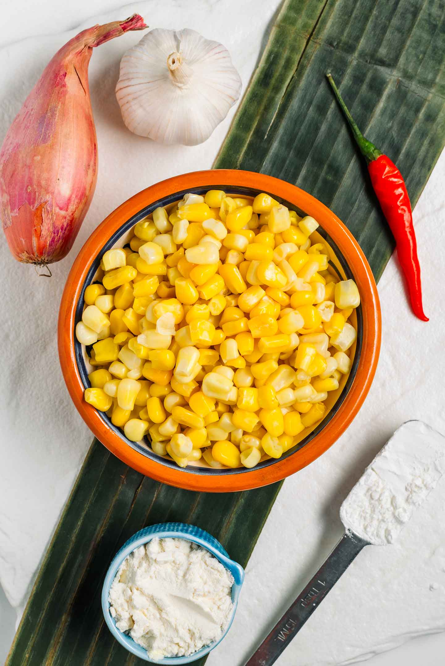 Top down view of ingredients for vegan Indonesian corn fritters. Corn kernels fill a small bowl surrounded by garlic, shallot, red chili, cornstarch, and flour.