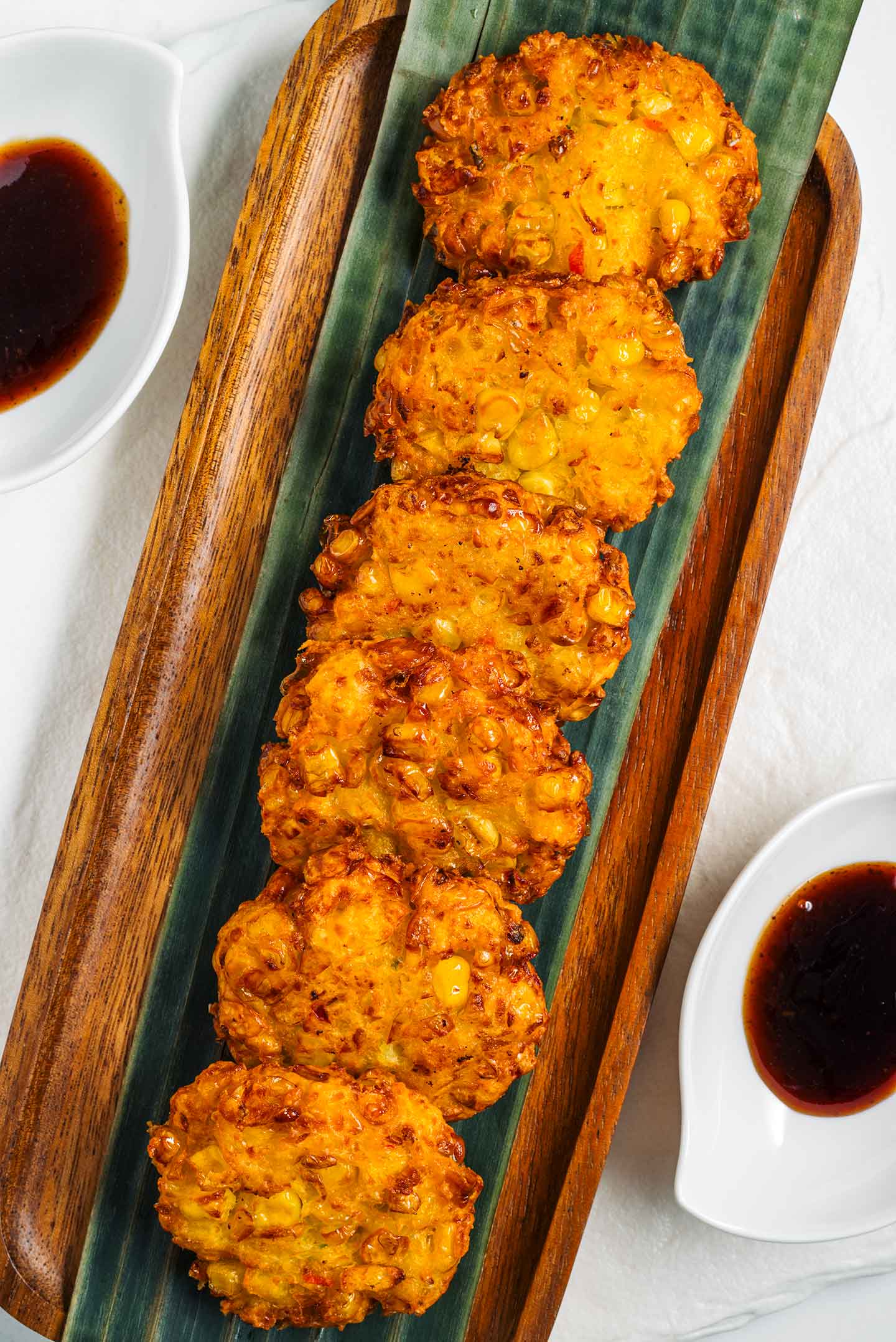 Top down view of vegan Indonesian corn fritters (perkedels) lined up on a tray. The fritters are round, golden, and pieces of caramelized sweet corn are visible. Dipping sauce rests beside the tray.