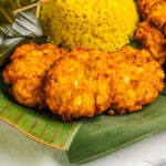 Close-up of three Indonesian perkedels on a platter lined with banana leaves. The vegan Indonesian corn fritters are golden and crispy.
