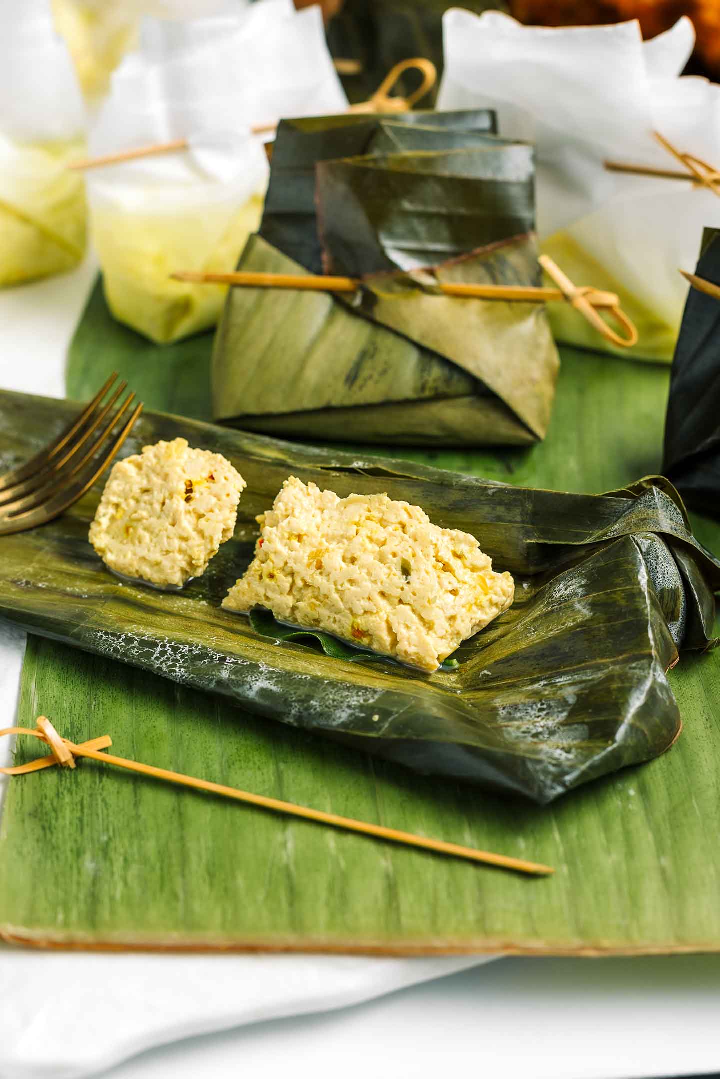 A steamed piece of tofu is displayed in the centre of an opened banana leaf steaming packet. The tofu is formed like a soft dumpling speckled with herbs and spices. Unopened tofu pockets stand in the background.