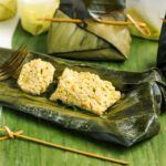 A steamed piece of tofu is displayed in the centre of an opened banana leaf steaming packet. The tofu is formed like a soft dumpling speckled with herbs and spices.