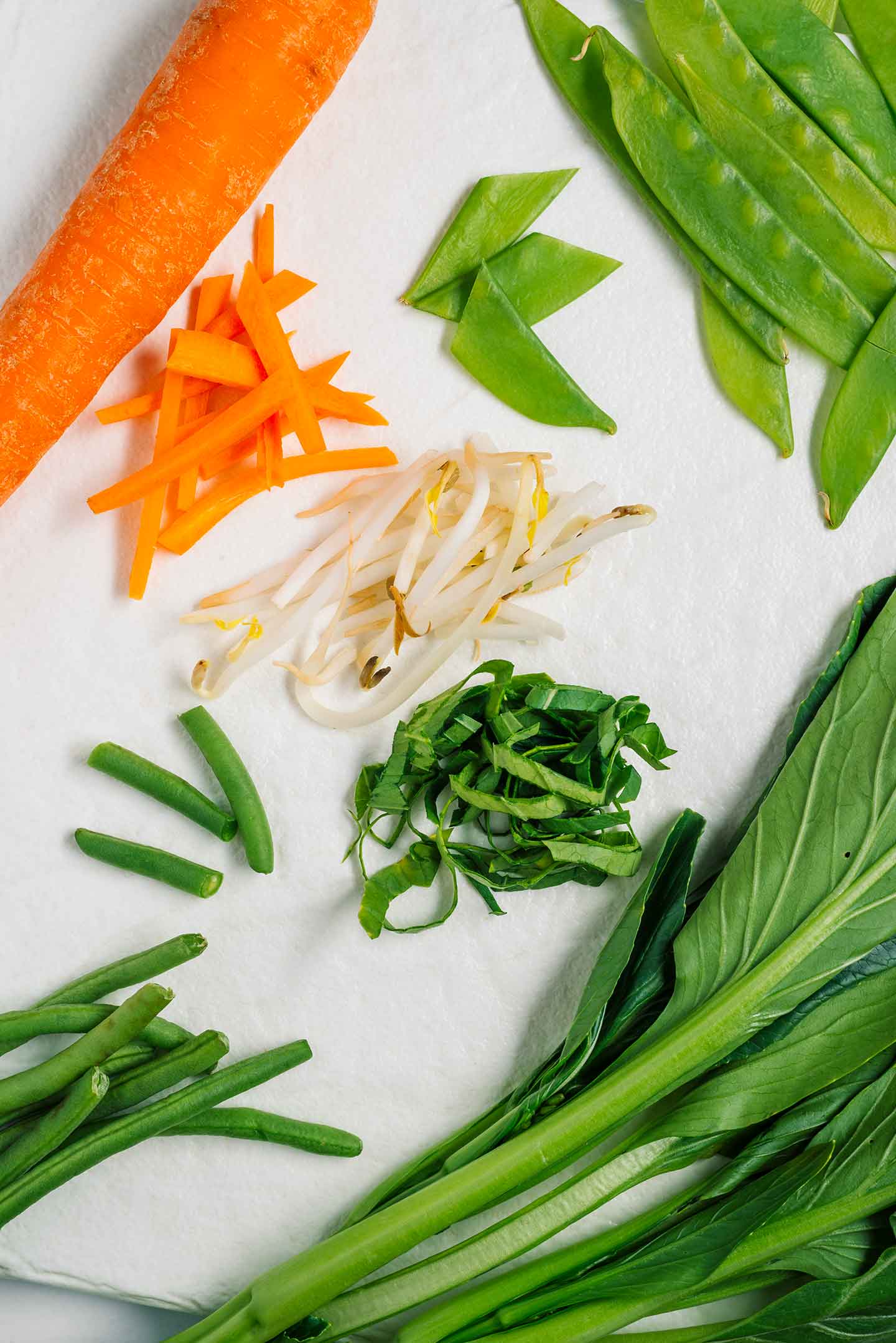 Top down view of chopped vegetables. A carrot is julienned, snow peas and green beans are cut in thirds, yu choy is ribboned and bean sprouts lay in the centre. 