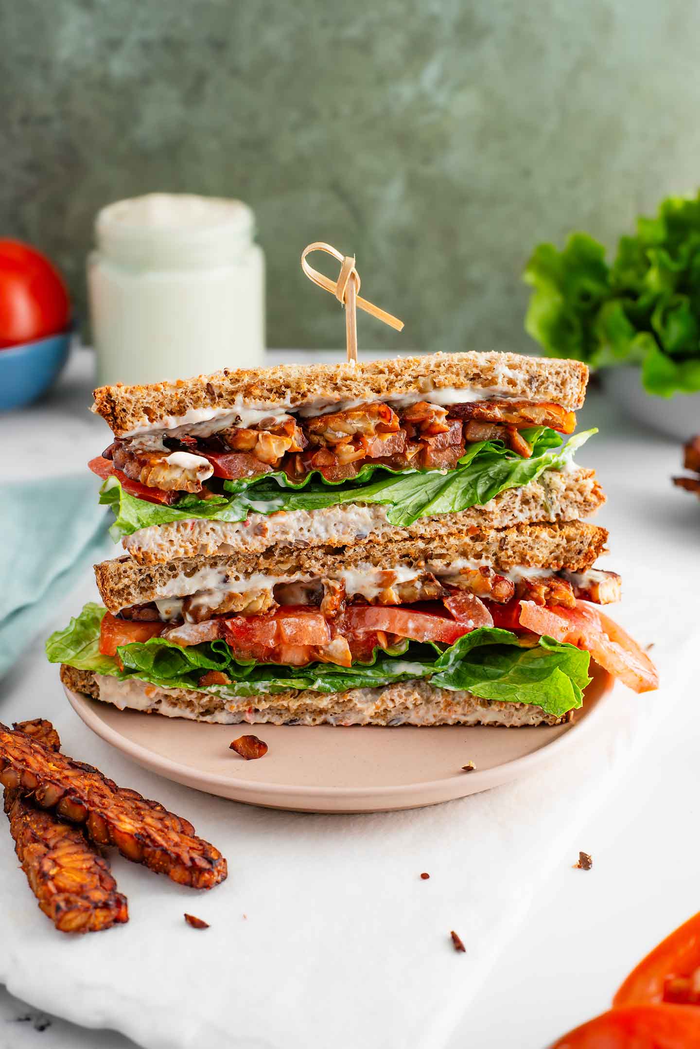 Two halves of a vegan BLT sandwich are stacked. Crispy tempeh bacon, lettuce, tomato, and oil-free mayonnaise are sandwiched between slices of seeded bread.
