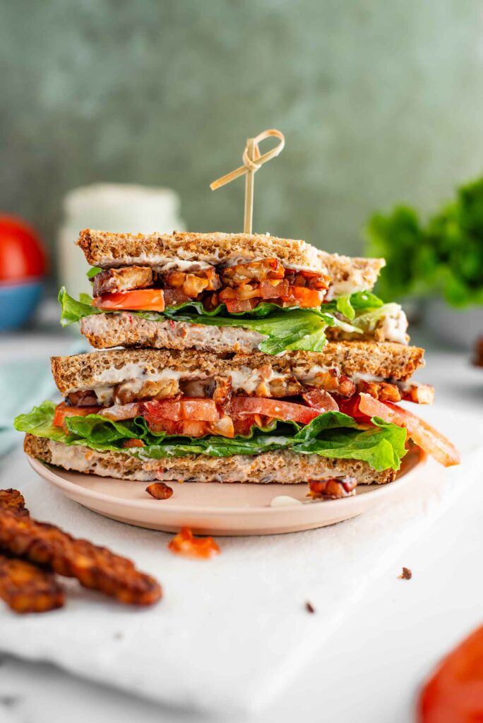 Side view of two halves of a vegan BLT stacked on one another. The top half has a large bite removed. Crispy tempeh bacon, lettuce, tomato, and oil-free mayonnaise fill the packed sandwich. Additional ingredients surround the sandwich.