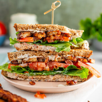 Side view of two halves of a vegan BLT stacked on one another. The top half has a large bite removed. Crispy tempeh bacon, lettuce, tomato, and oil-free mayonnaise fill the packed sandwich.