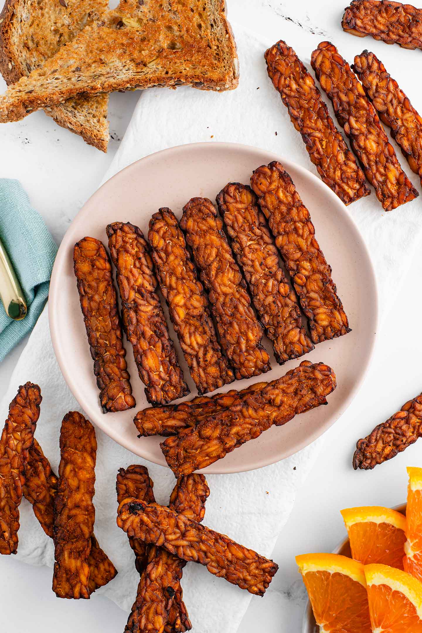 Slices of crispy tempeh bacon fill a plate. The tempeh is dark brown with charred edges. More bacon surrounds along with toast and orange slices. 