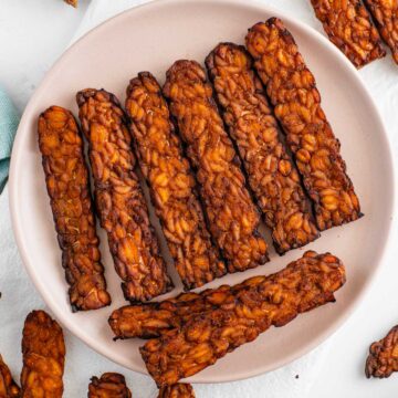 Slices of crispy tempeh bacon fill a plate. The tempeh is dark brown with charred edges.