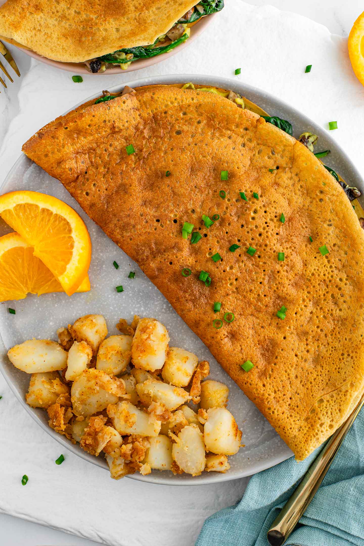 Top down view of a golden vegan omelette made from chickpea flour. The fillings peak out from the side and chives garnish the top. Home fries and orange slices garnish the plate.
