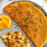 Top down view of a golden vegan omelette made from chickpea flour. Home fries and orange slices garnish the plate.